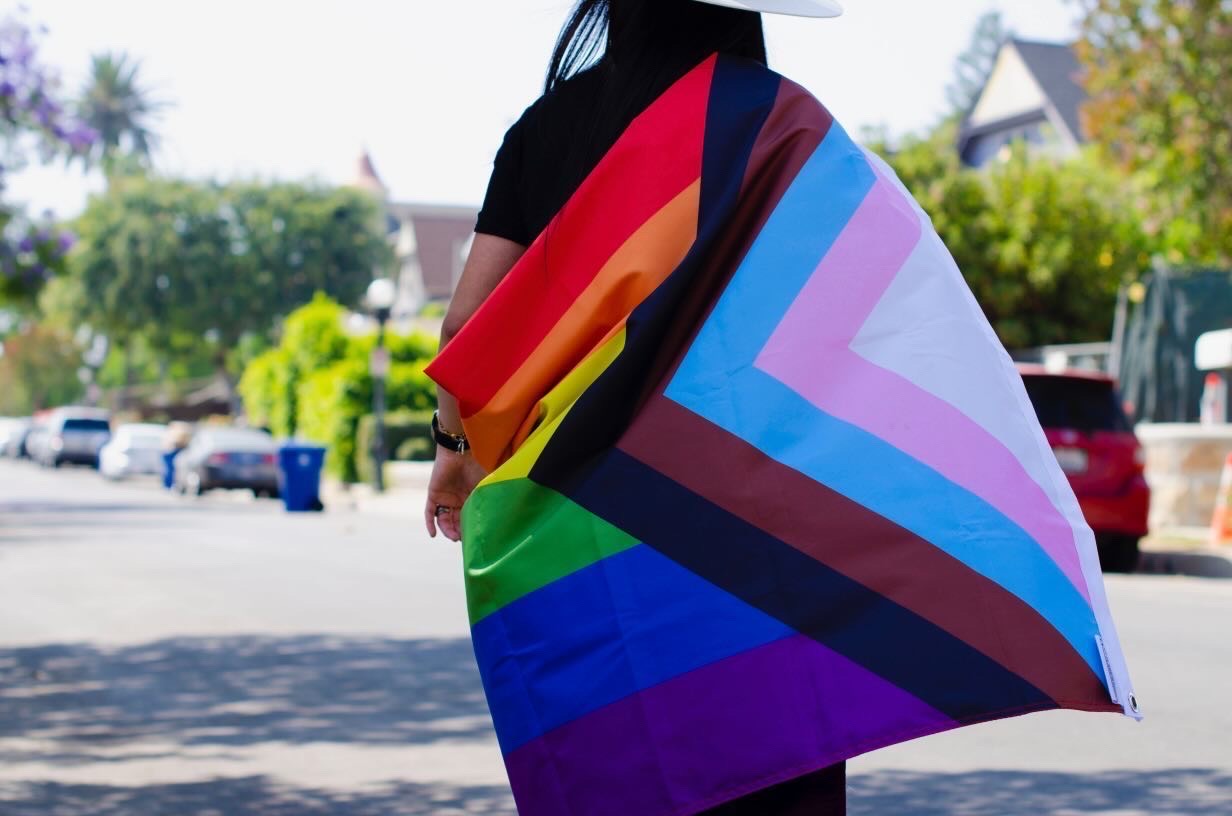 A person with a Progress Pride Flag wrapped around them as they walk down a street.