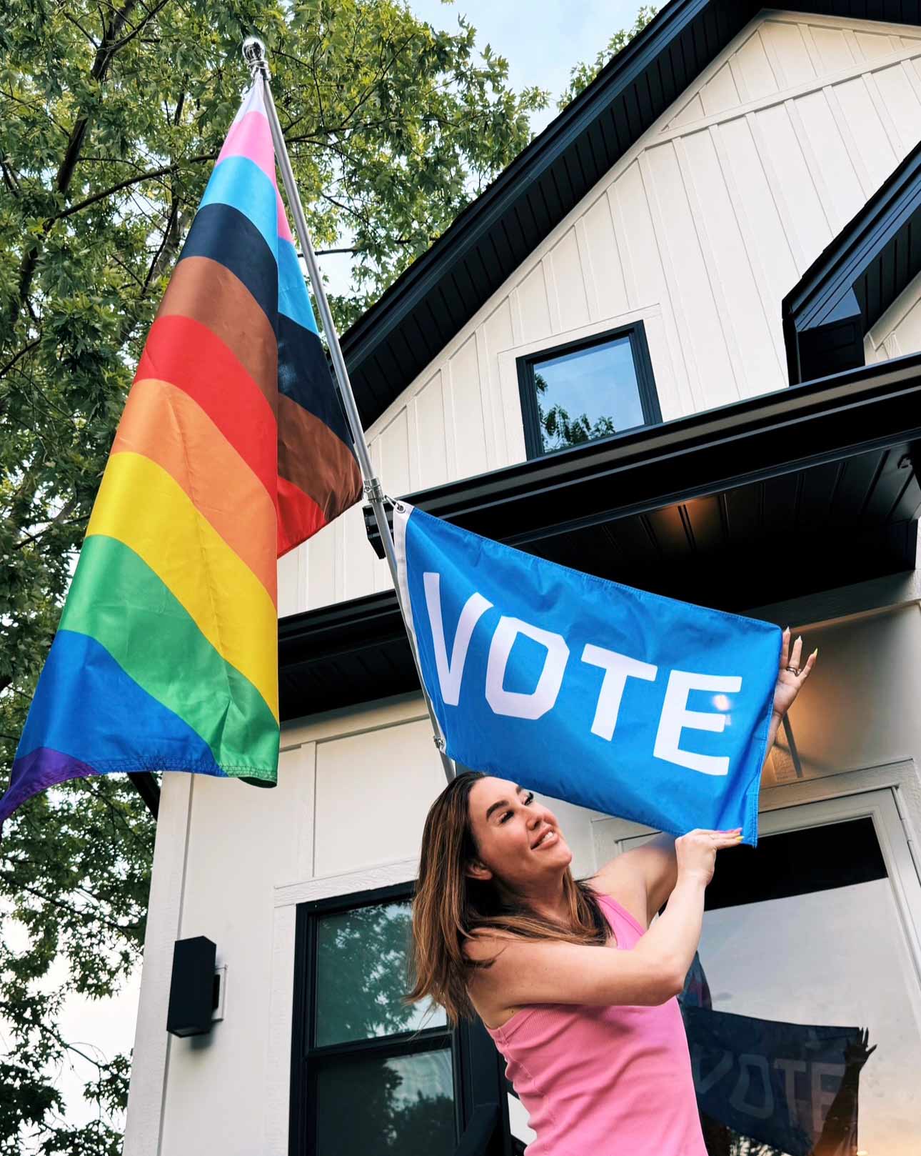 New 11-stripe diagonal LGBTQ Pride flag flying on a pole with a smaller blue VOTE flag being held by someone in front of their house.