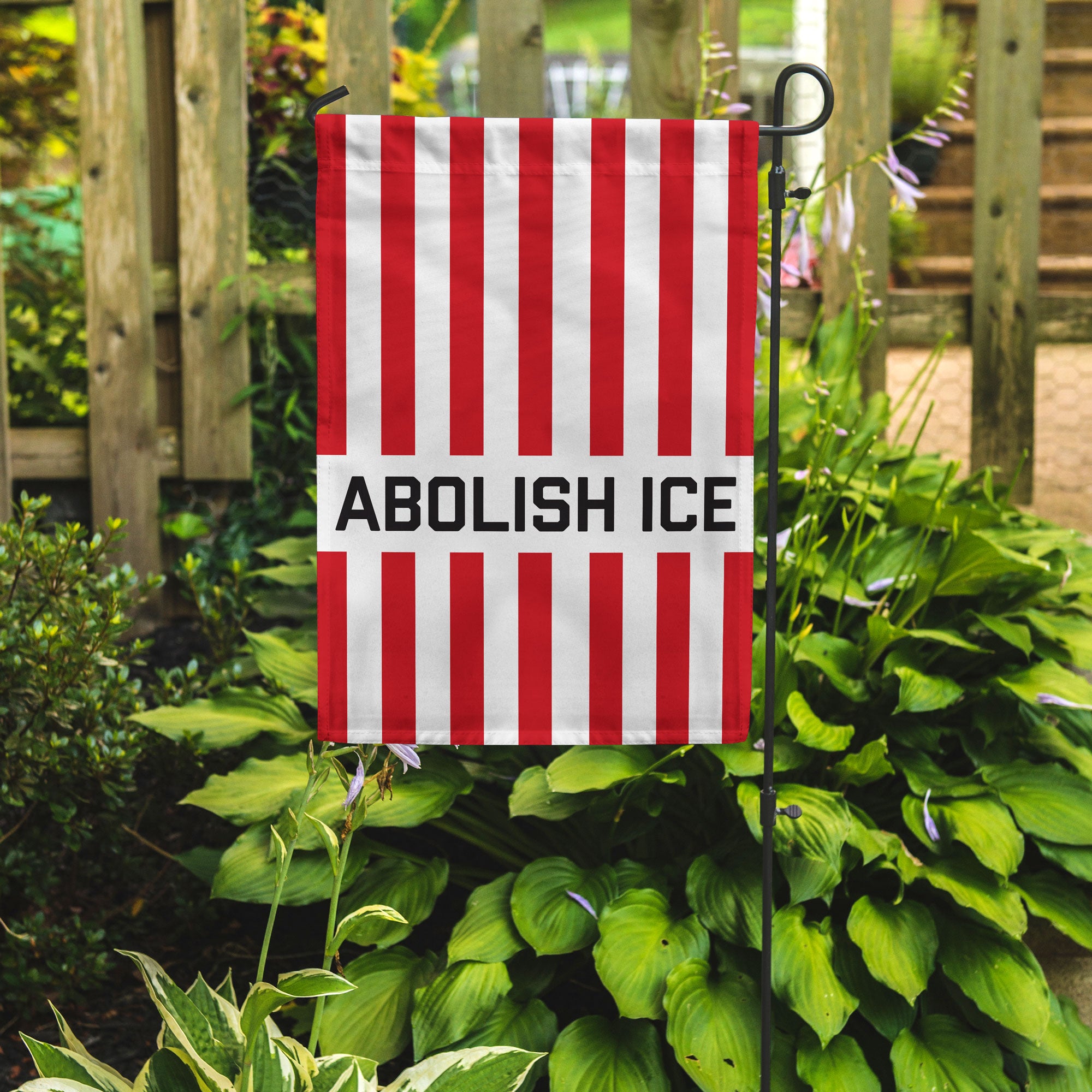 Red and white vertical striped garden flag with 'ABOLISH ICE' text in a garden setting