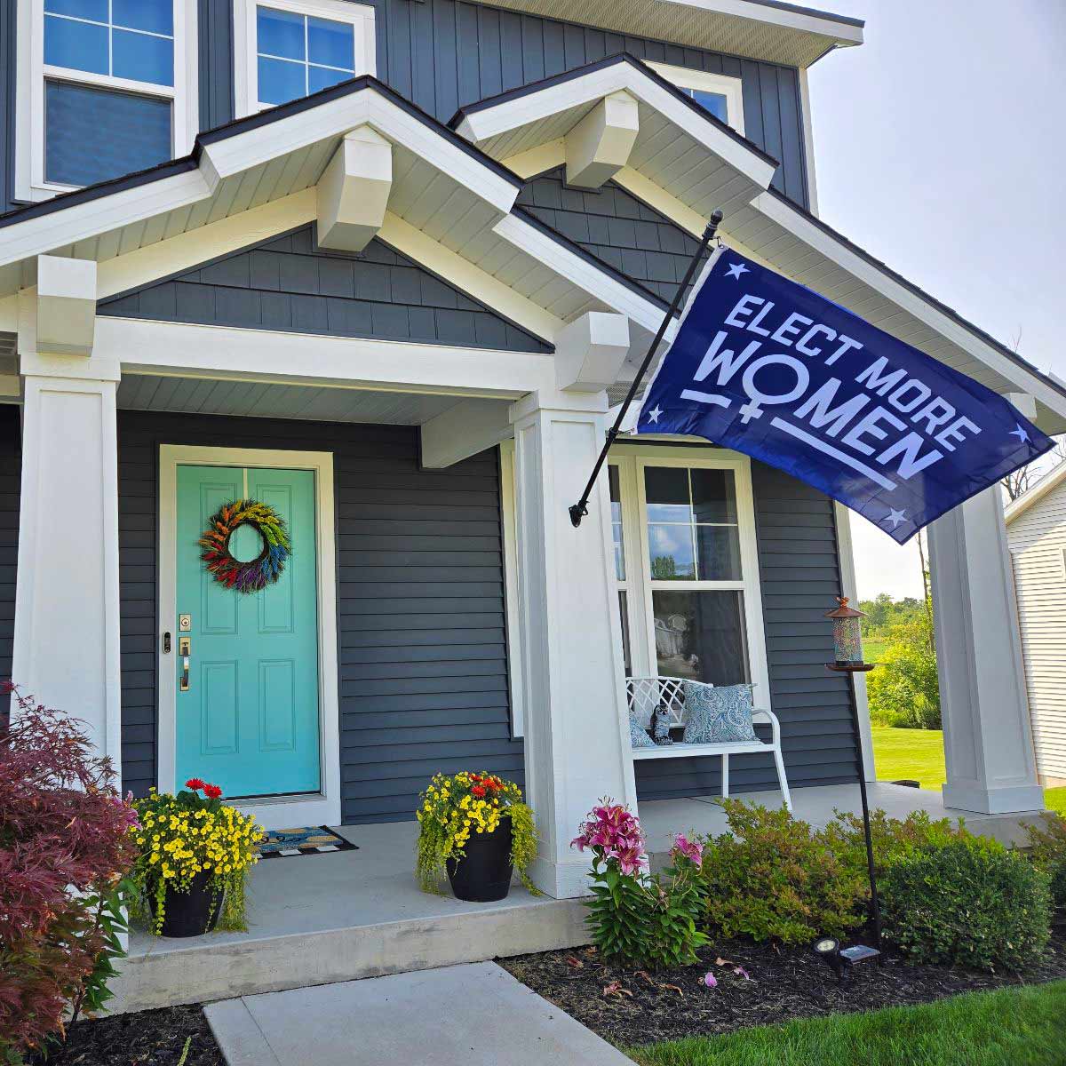 a blue flag with the words elect more women in the center with four stars and the international symbol for women flying on a flagpole in front of an lgbtq+ pride flag