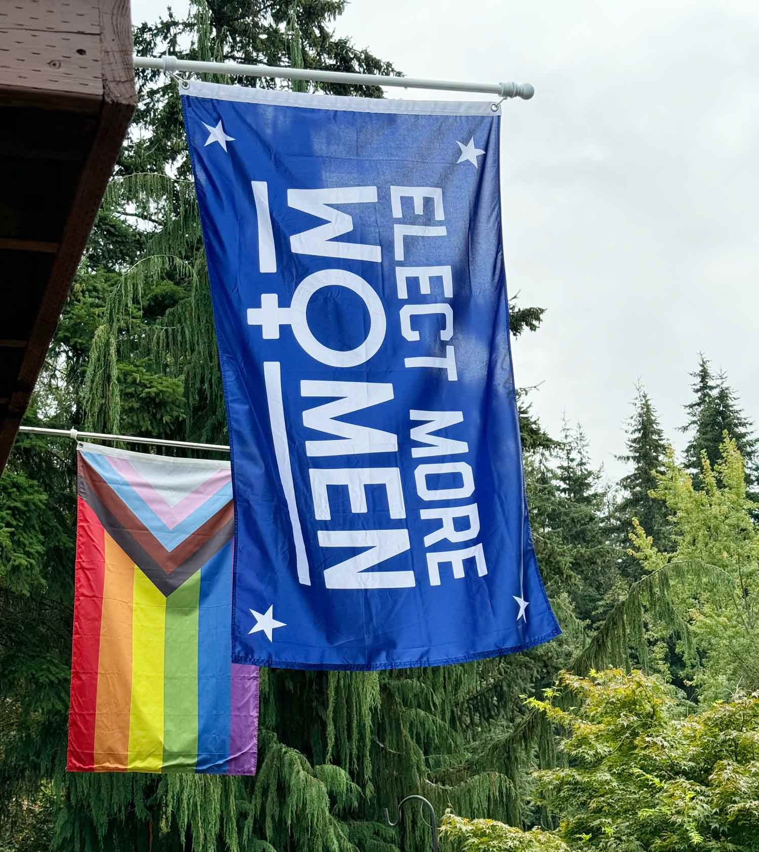 a blue flag with the words elect more women in the center with four stars and the international symbol for women flying on a flagpole in front of an lgbtq+ pride flag