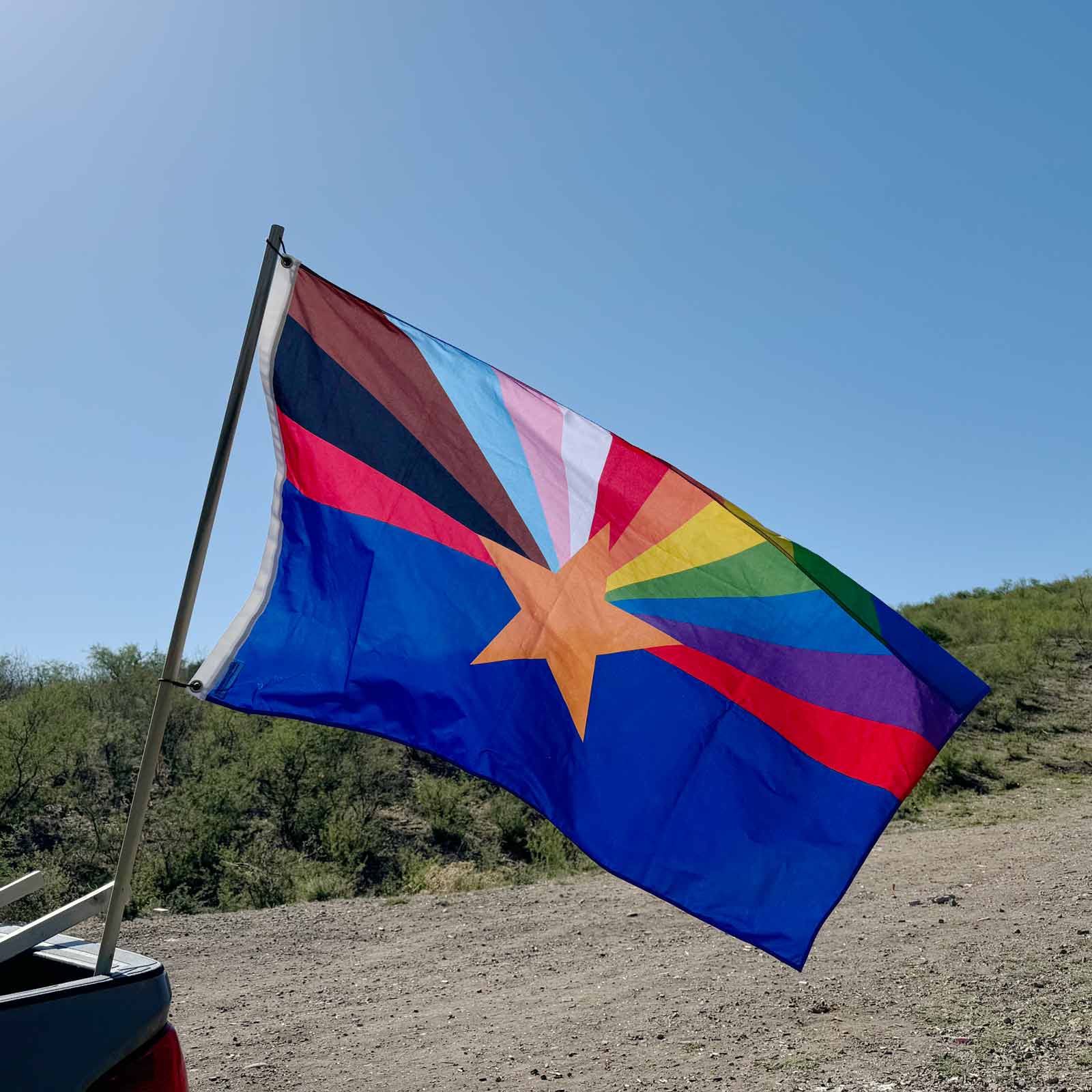 Arizona state flag with the rays replaced with an LGBTQ+ Rainbow flying behind a truck in the desert