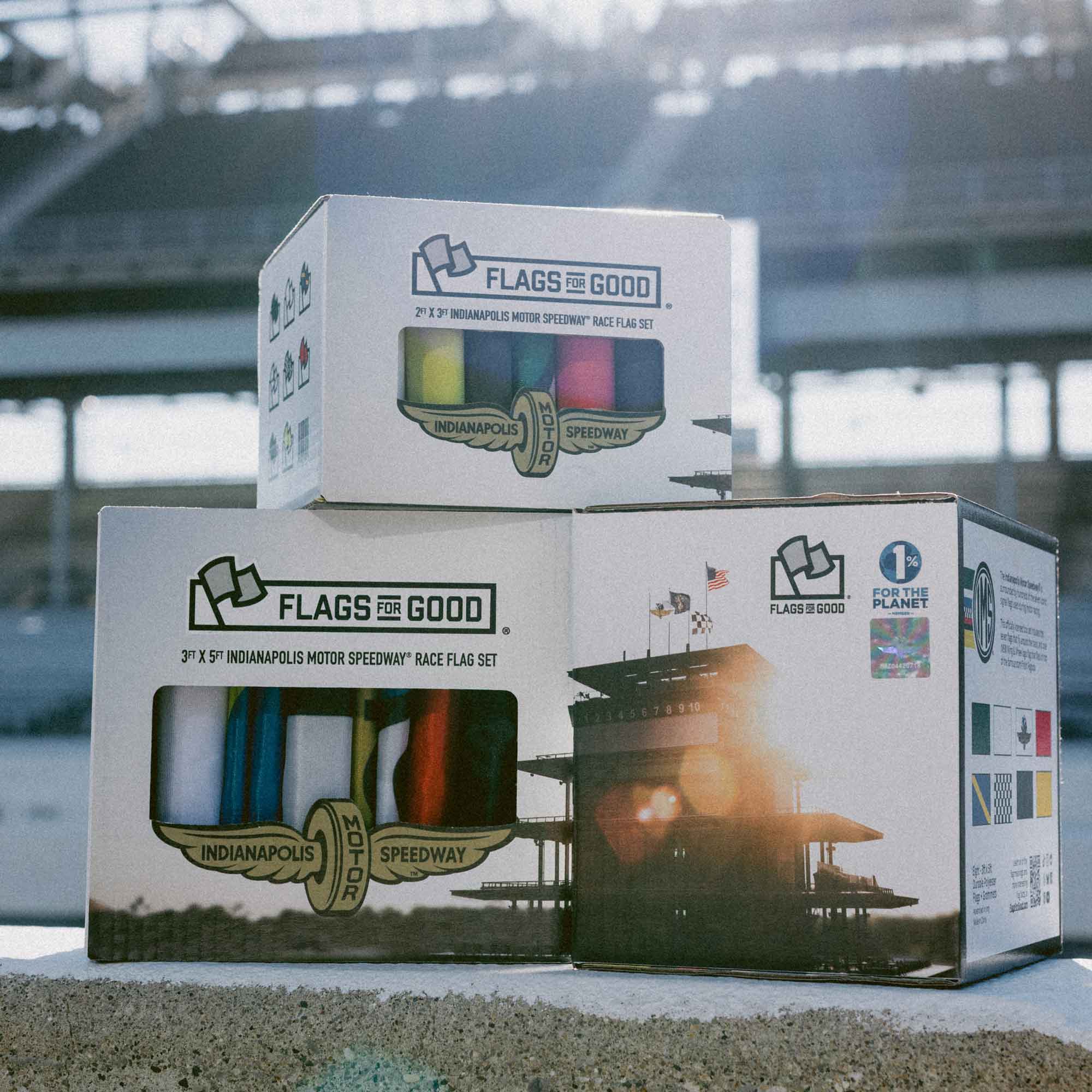 Three boxes of 'Flags for Good' racing flags box sets on a concrete surface with a blurred background of the IMS