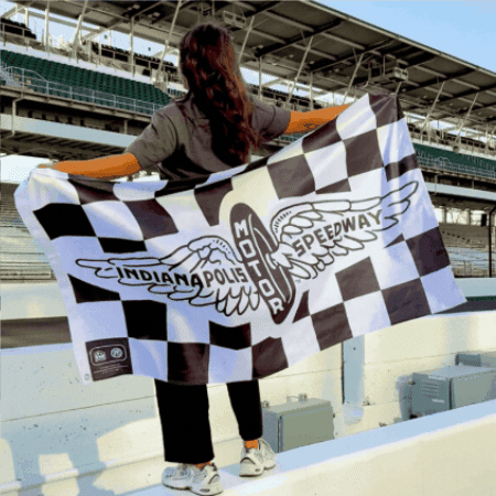 Woman standing on a race track wall with a checkered flag with vintage IMS logo, wearing a grey shirt and black pants.