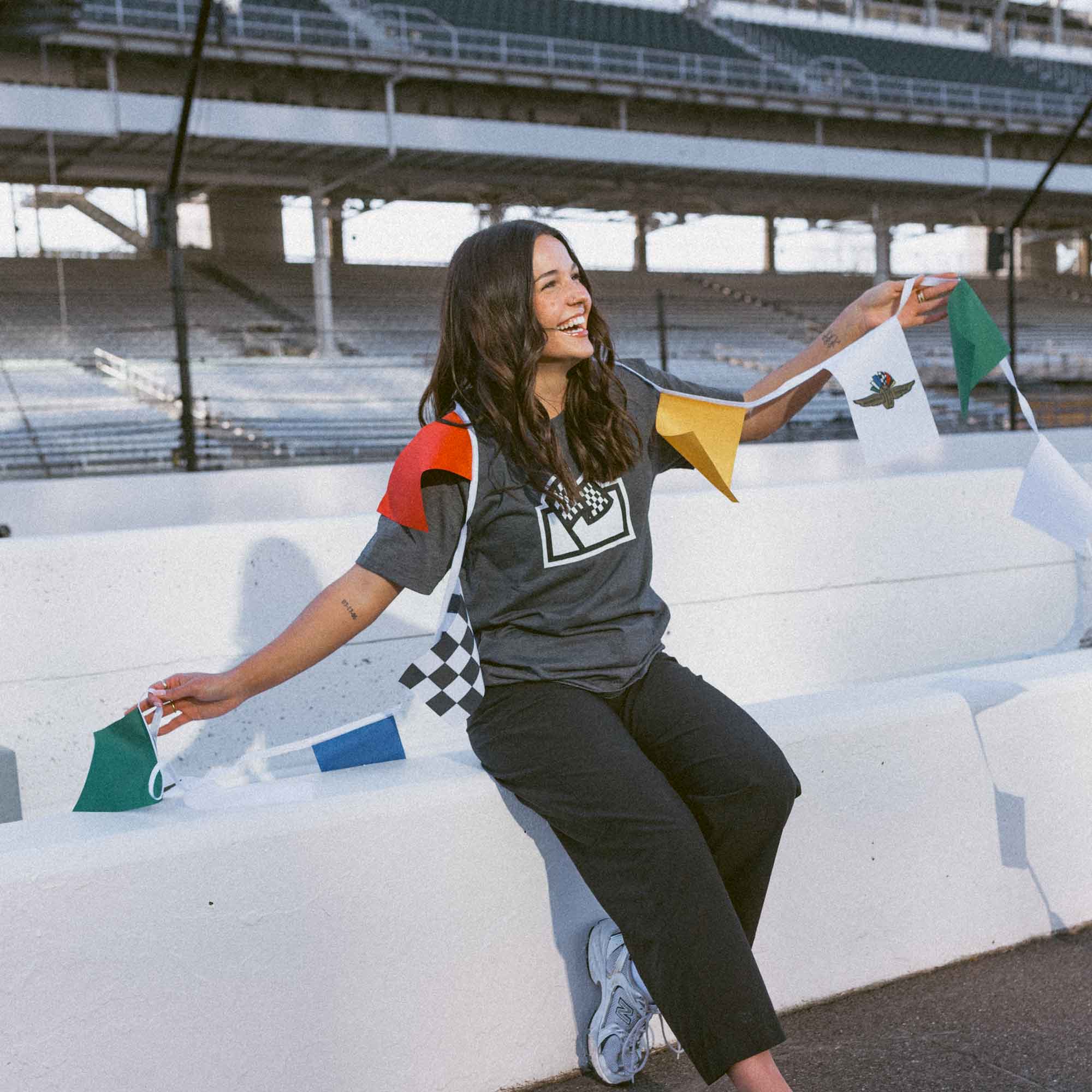 Woman sitting on a race track wall with race flags bunting, wearing a Flags For Good shirt and black pants.