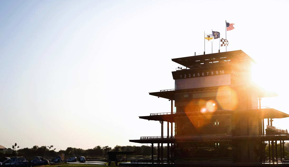 Silhouette of the Indianapolis Motor Speedway race track control tower with flags against a sunset sky.