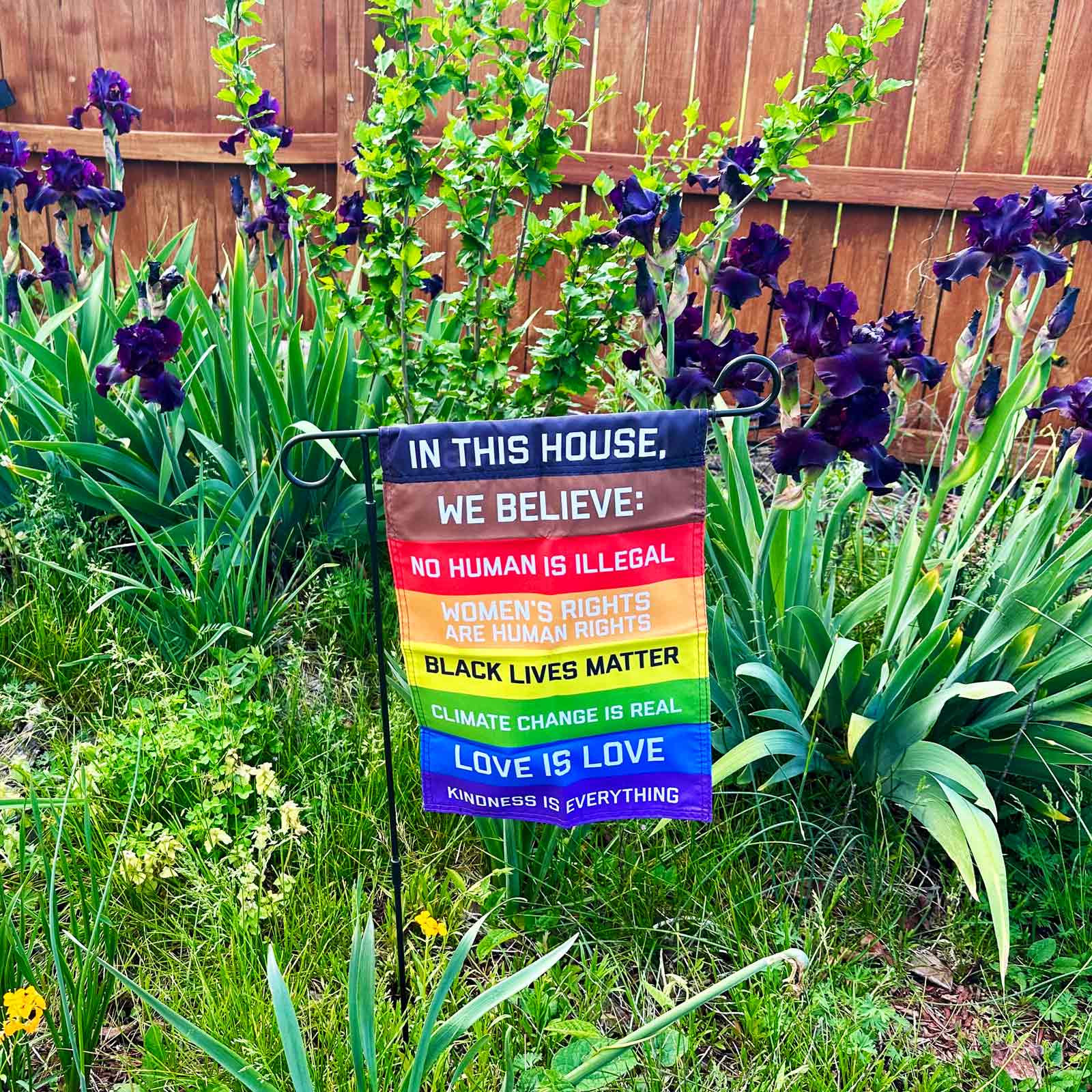 "In this House We Believe: No human is Illegal, Women's RIghts are Human Rights, Black Lives Matter, Cliamate Change is Real, Love is Love, Kindness is everything" garden flag hanging on a stand in a garden