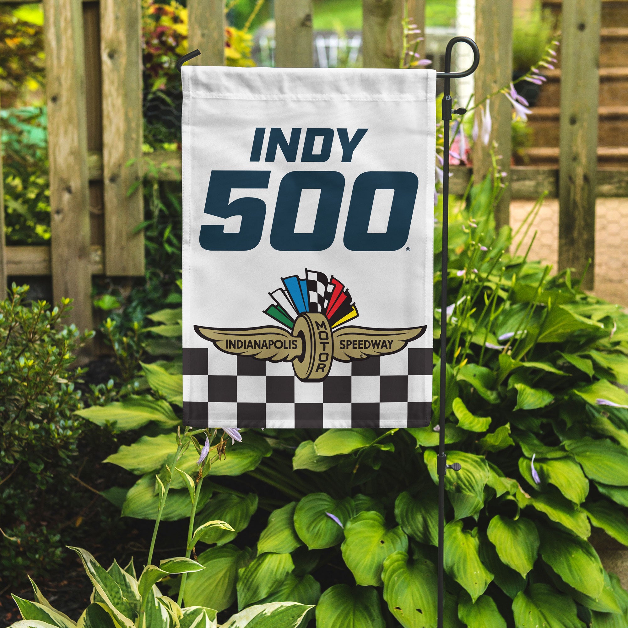 "Indy 500" in blue on a white garden flag with an Indianapolis Motor Speedway logo on a checkered flag on the bottom in a garden
