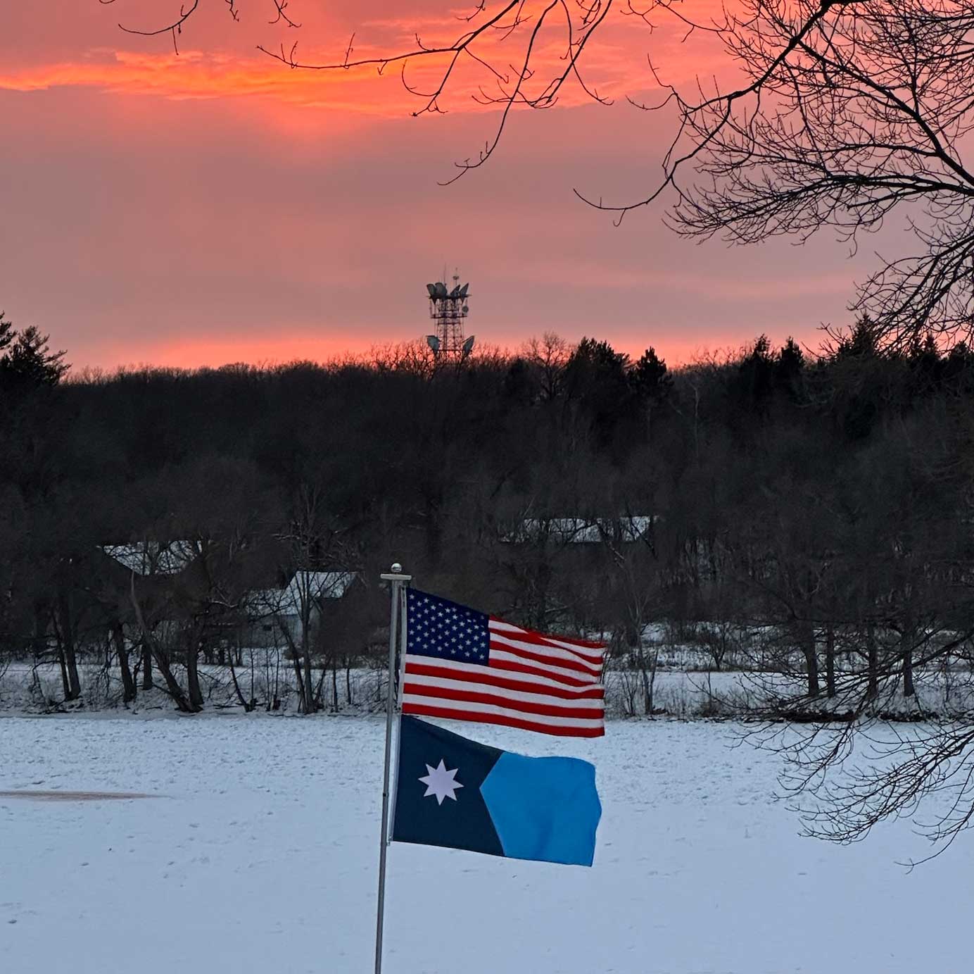 New Updated Minnesota State Flag flying on a flag pole with an American flag in front of a sunset.