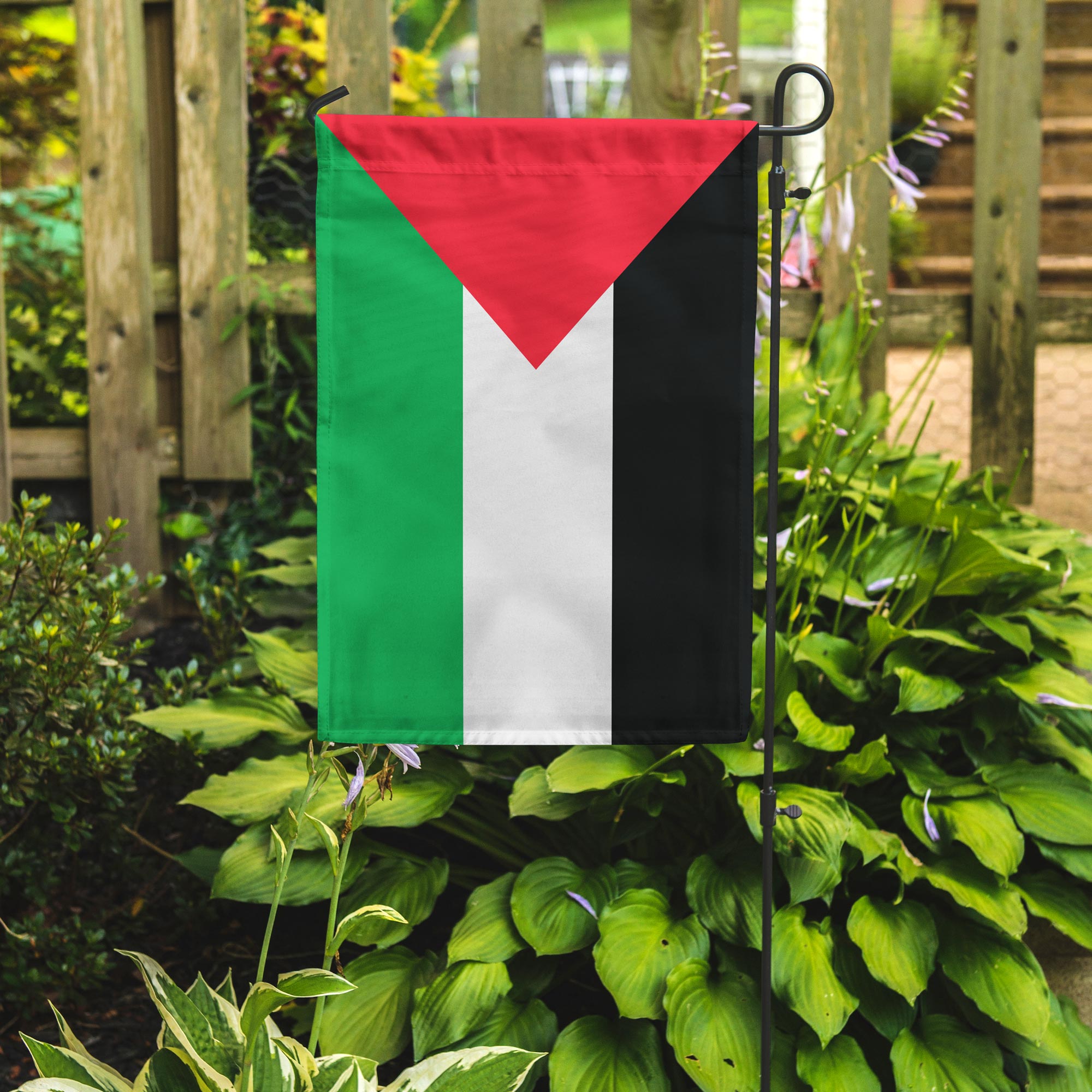 Palestine flag with red triangle on white, green, and black stripes on a garden flagpole surrounded by green plants.