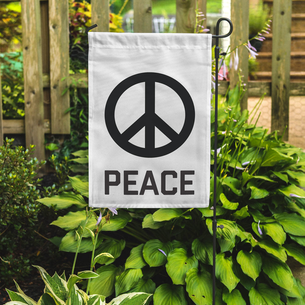 A white rectangular garden flag with a peace sign and the word 'PEACE' printed below it, mounted on a metal pole.