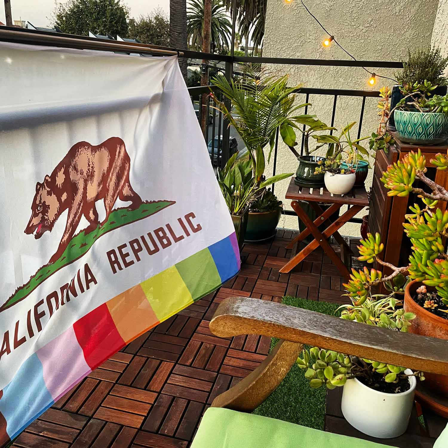 California state flag with a rainbow on the bottom instead of the red stripe hanging on a balcony with house plants