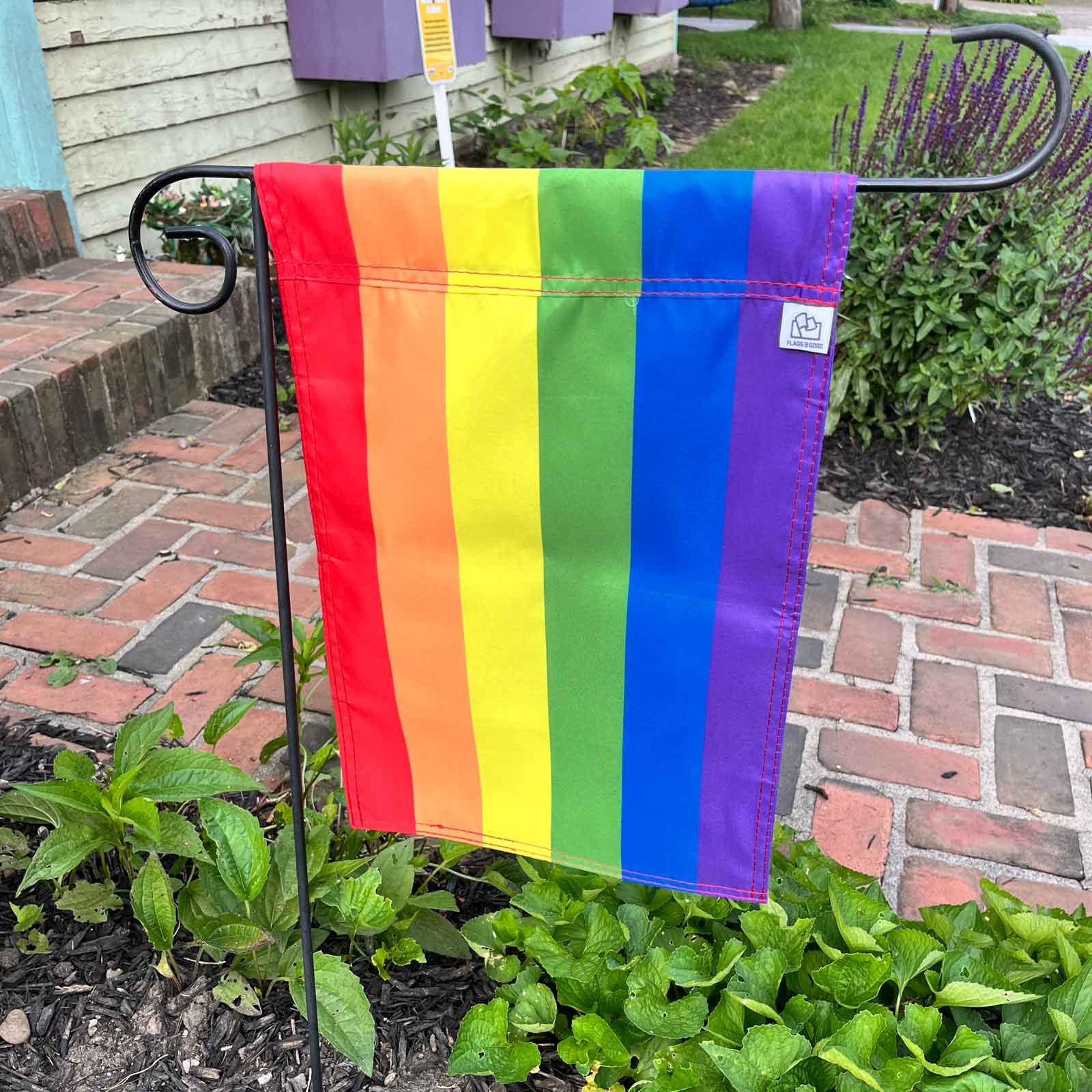 6-Stripe Rainbow LGBTQ+ Pride Garden Flag by Flags For Good in a garden showing the tag