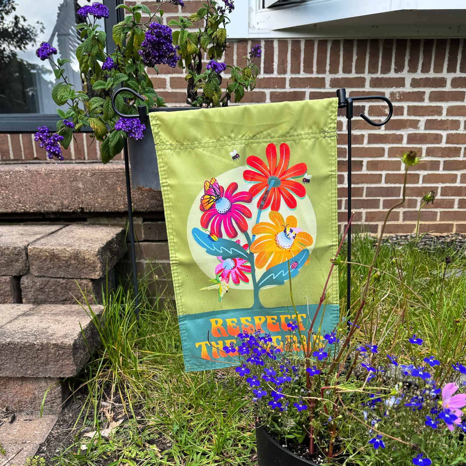 "Respect the Locals" garden flag with image of native plants, bees, and butterflies on a green background in front of a house