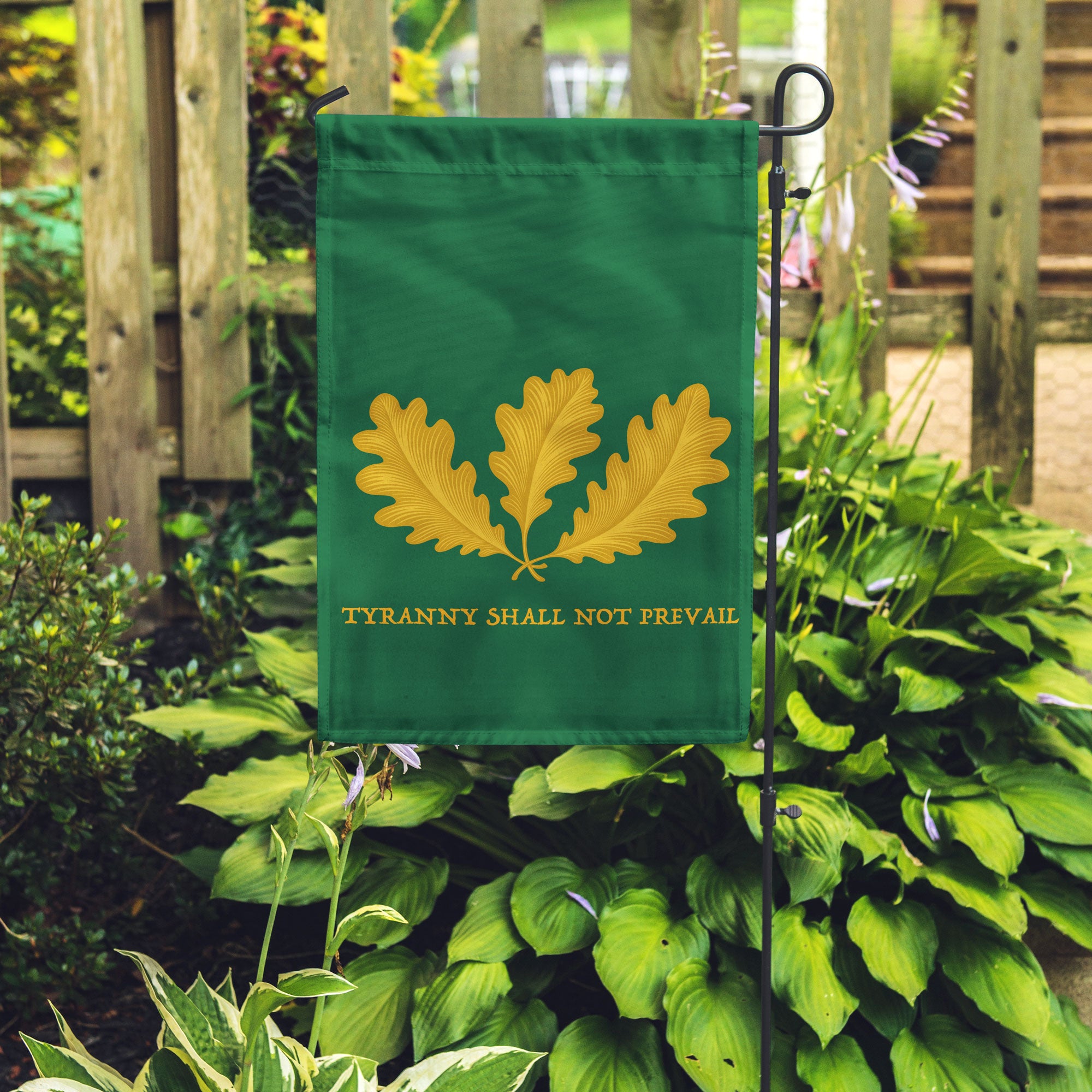 Green garden flag with yellow leaf design and text 'Tyranny Shall Not Prevail' in a garden setting.