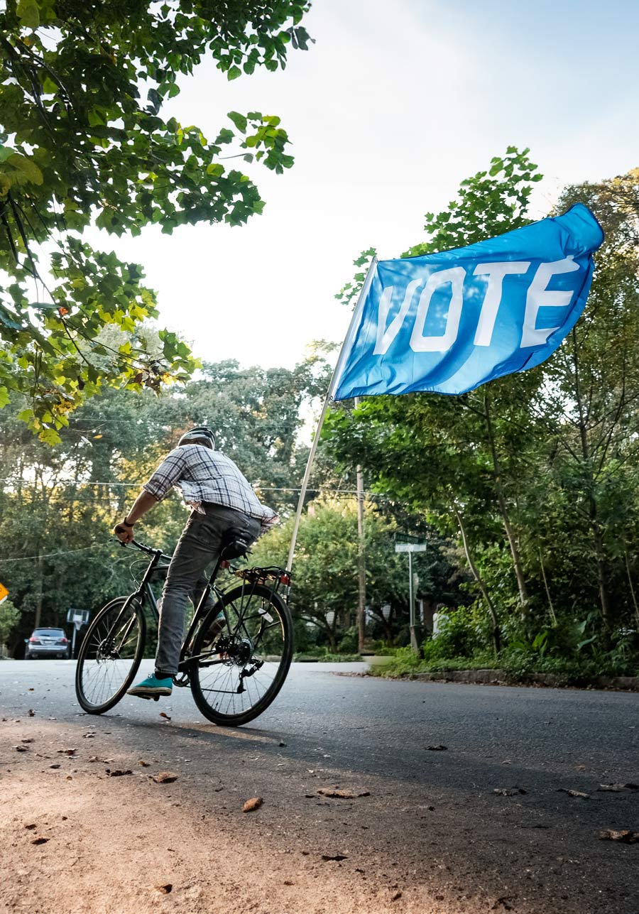 Biker with a blue "Vote" flag flying behind his bike riding down a street
