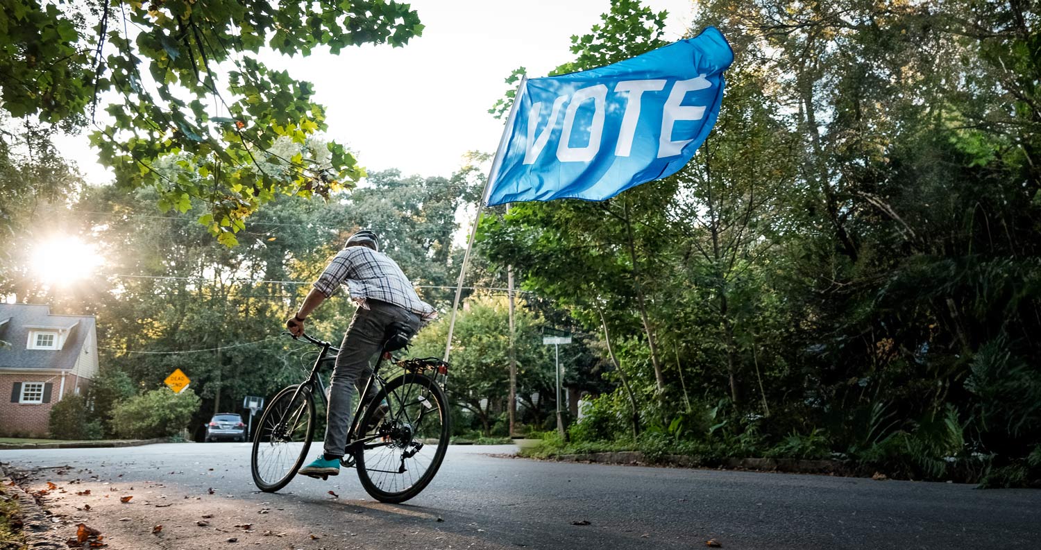 Biker with a blue "Vote" flag flying behind his bike riding down a street