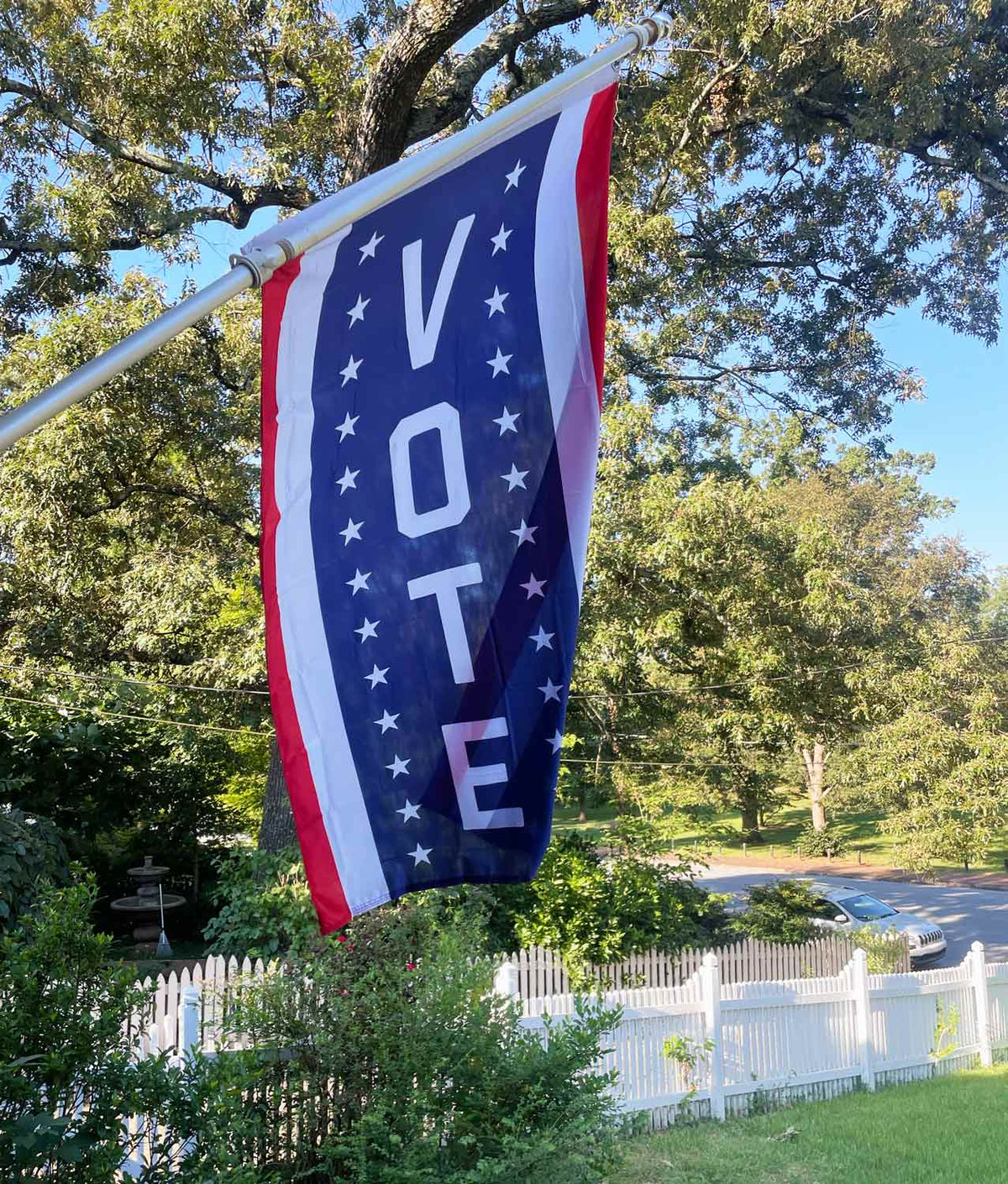 Vertical flag with red and white stripes on the sides and in the center, the word VOTE flanked with stars on blue flying on a flag pole in someone's yard