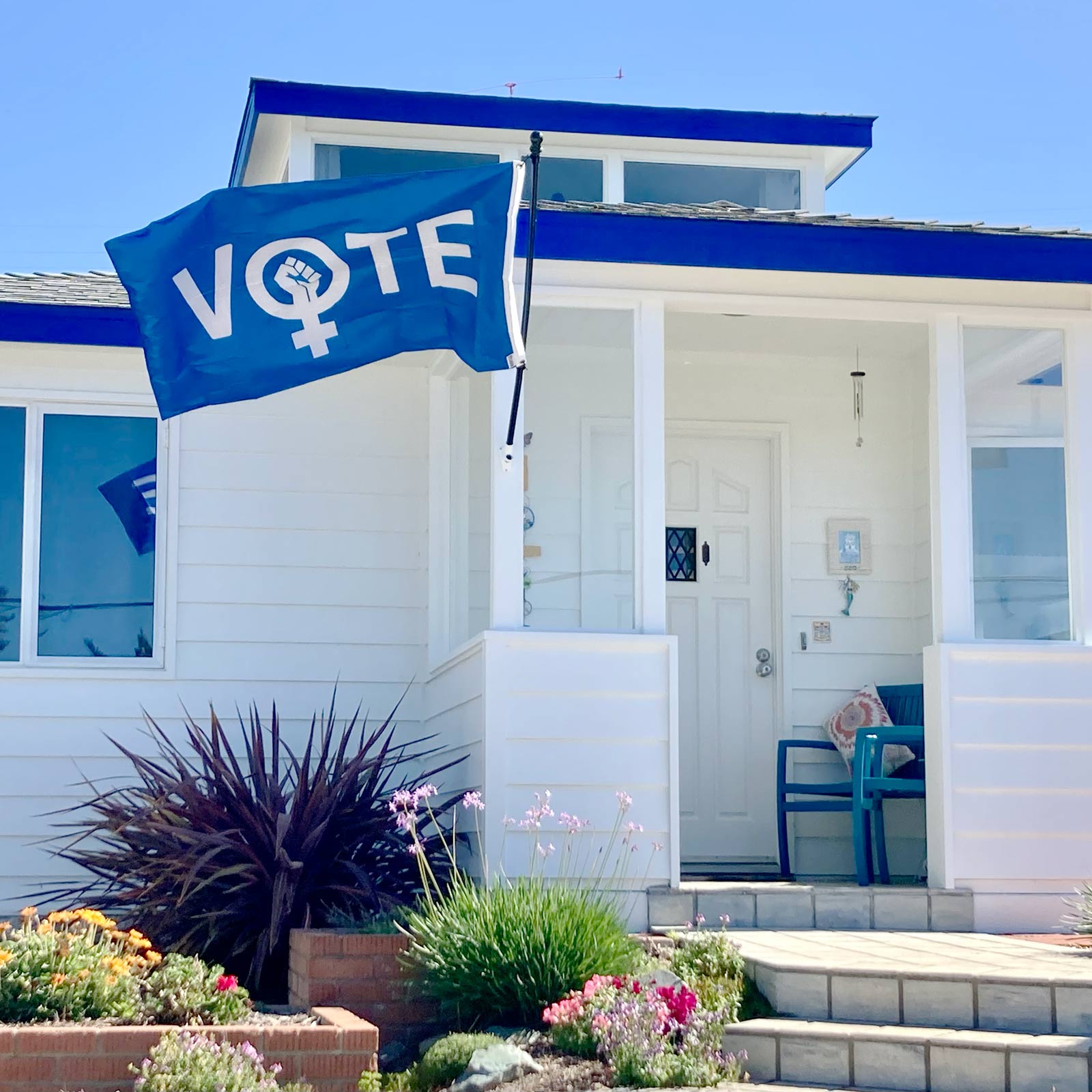 A blue flag with the word VOTE but the “O” is the symbol for females with a raised fist inside of it on a pole in front of a white house.