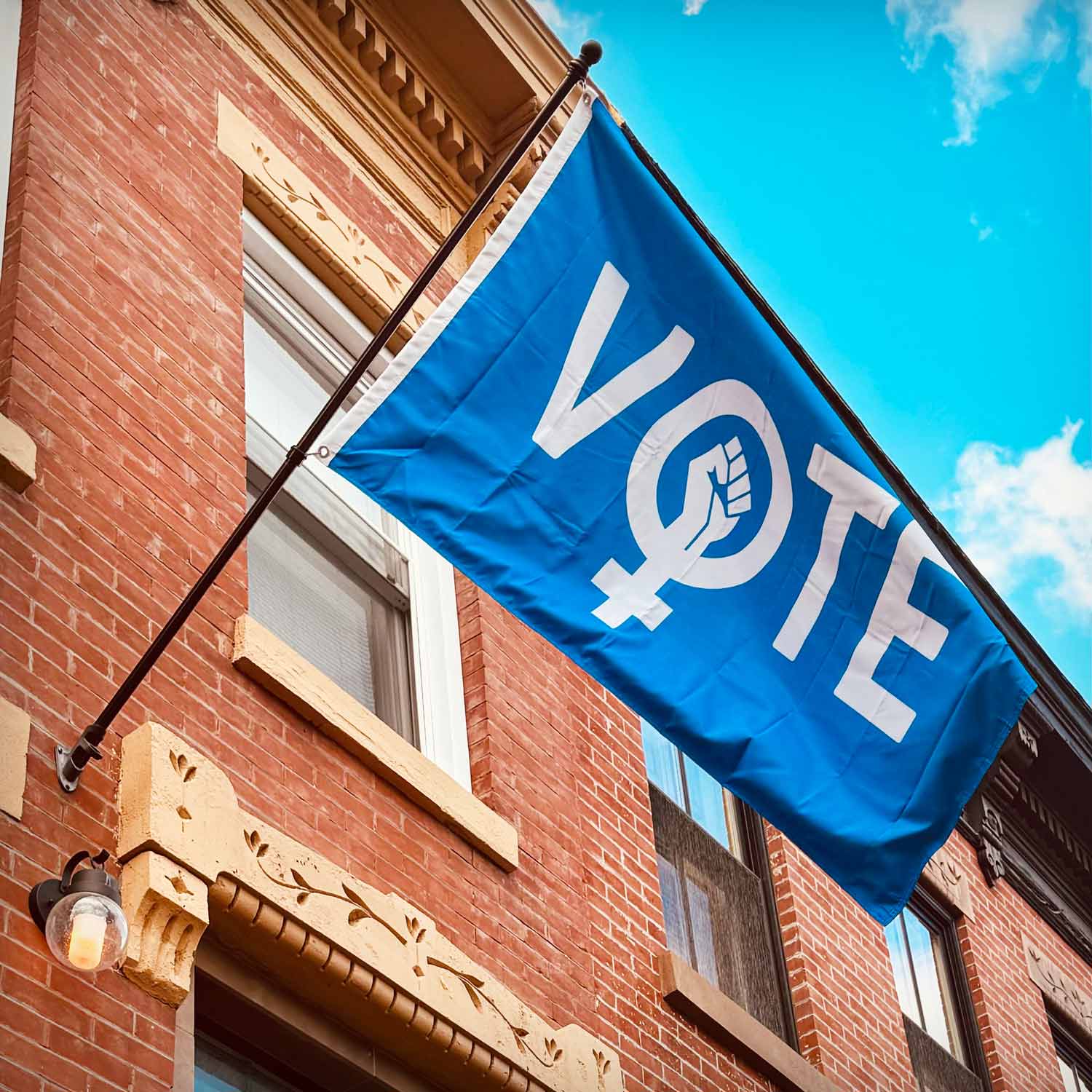 A blue flag with the word VOTE but the “O” is the symbol for females with a raised fist inside of it hanging from a flag pole in front of a brick building