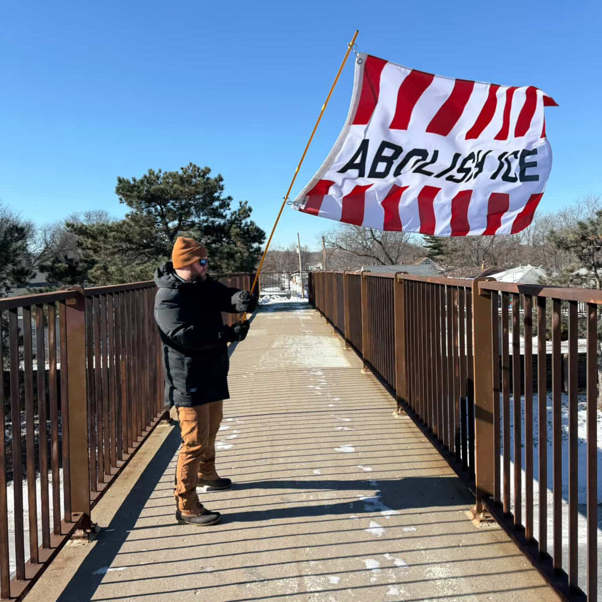 Person holding a red and white striped flag with the text "ABOLISH ICE" on a wooden bridge