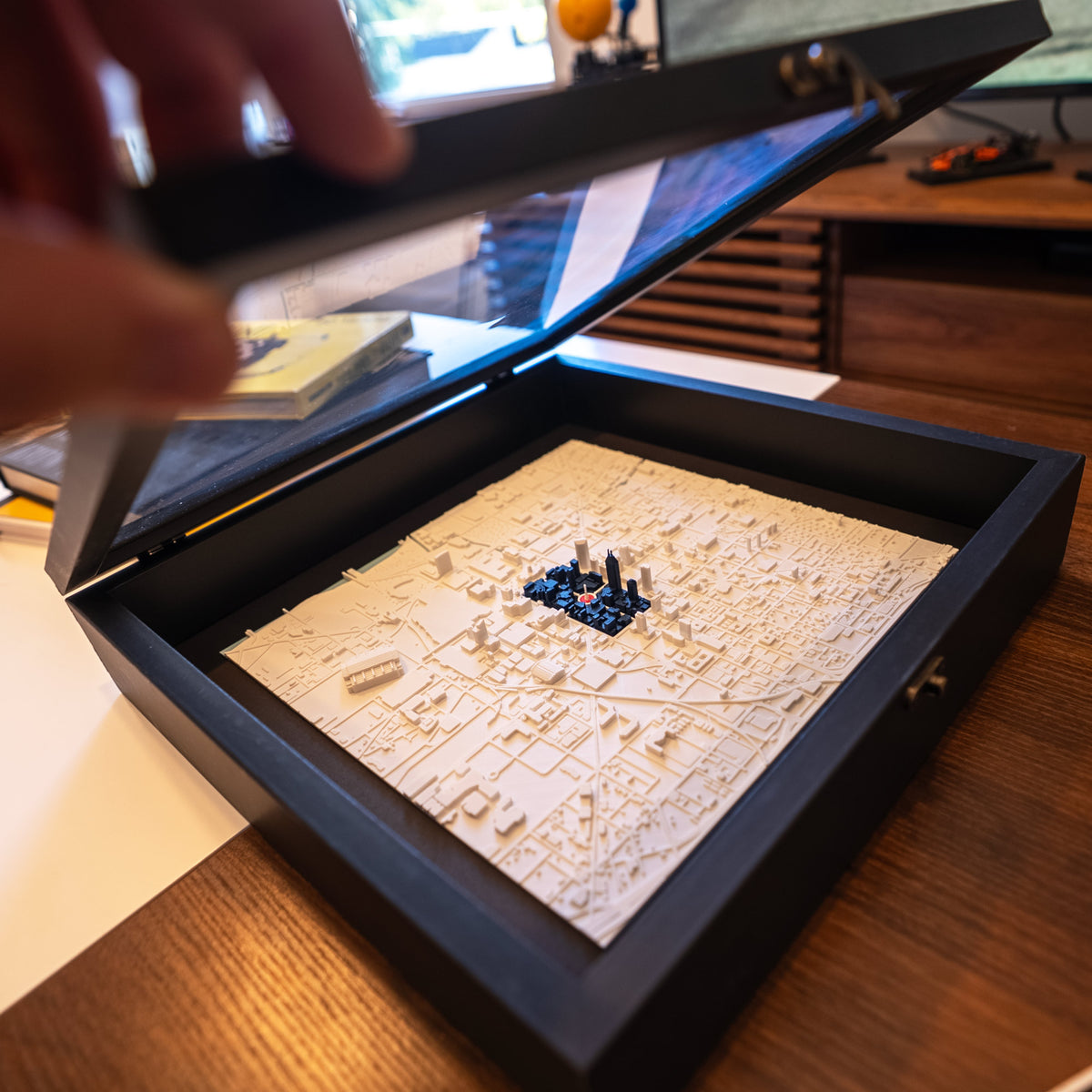 A hand is lifting the lid of a black shadow box frame that contains a 3D Indianapolis city model with the Indy flag portrayed by blue buildings and red on monument circle, set on a wooden desk.