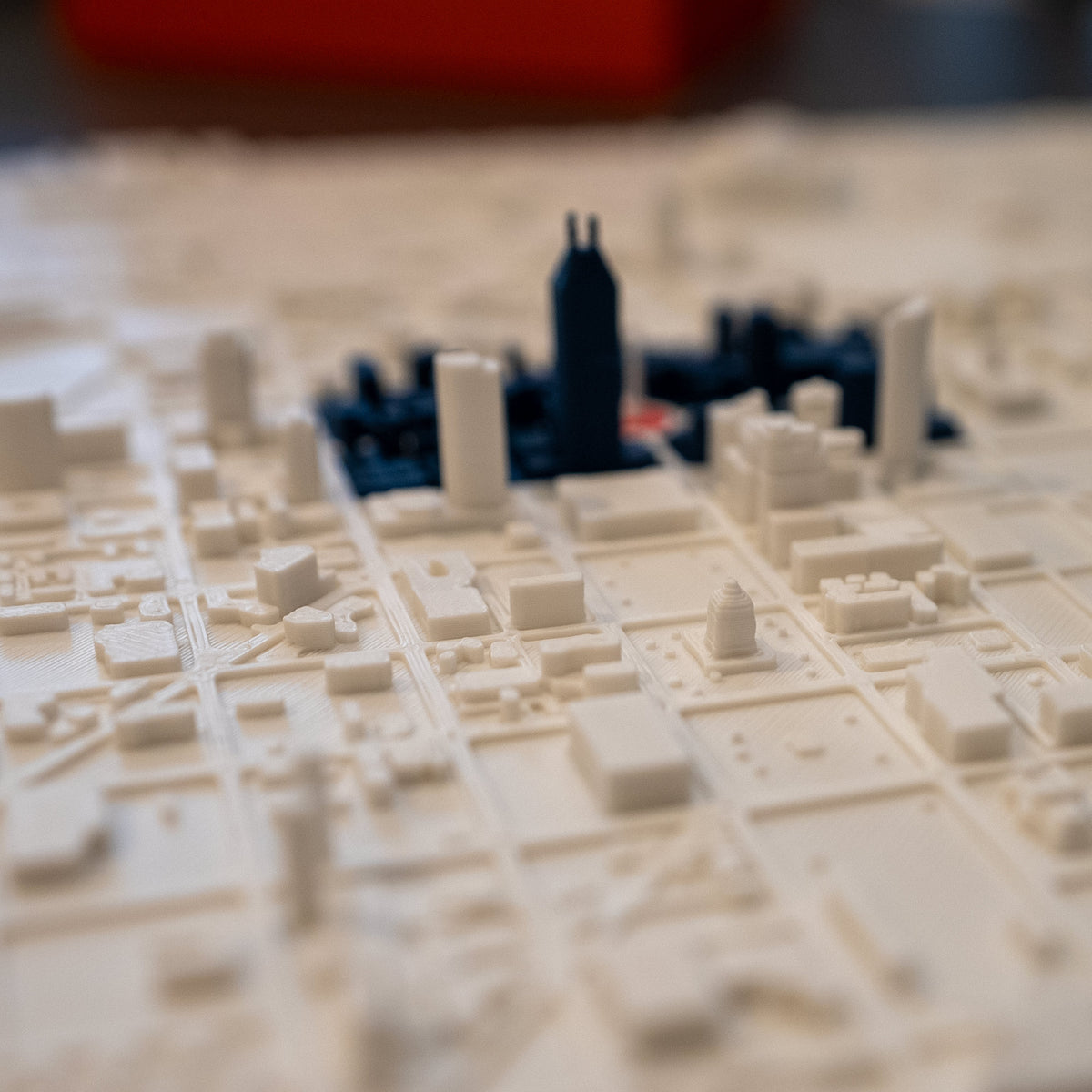 A close-up of a 3D-printed Indianapolis city map model with cream-colored street blocks and buildings, featuring the Indiana War Memorial in focus with downtown in blue and red (showing the indy flag) blurry in the background.