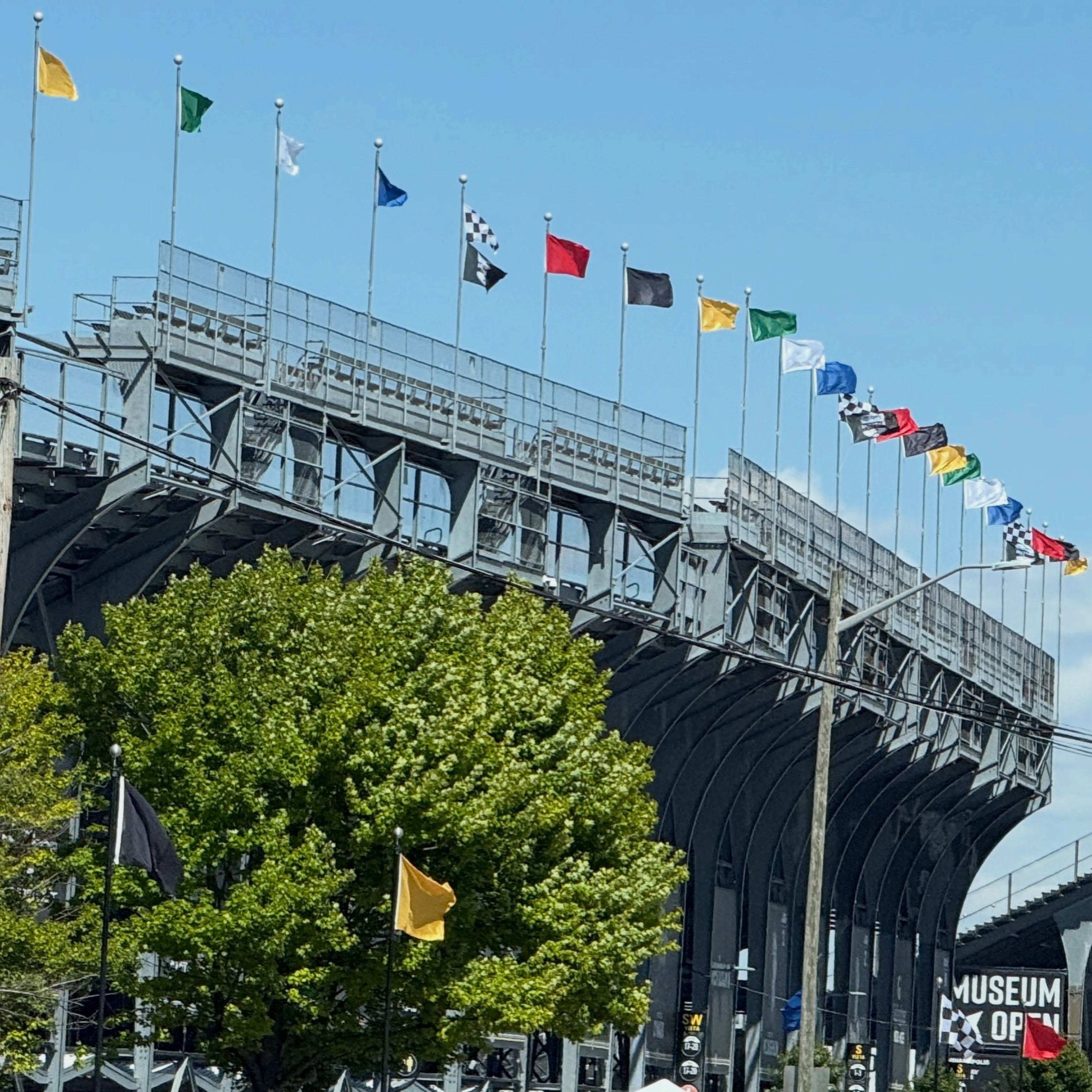 An outdoor architectural scene showing a large gray stadium or bridge structure under a clear blue sky, with multiple colorful flags along the top, green trees in the foreground, and a small white tent near the bottom.