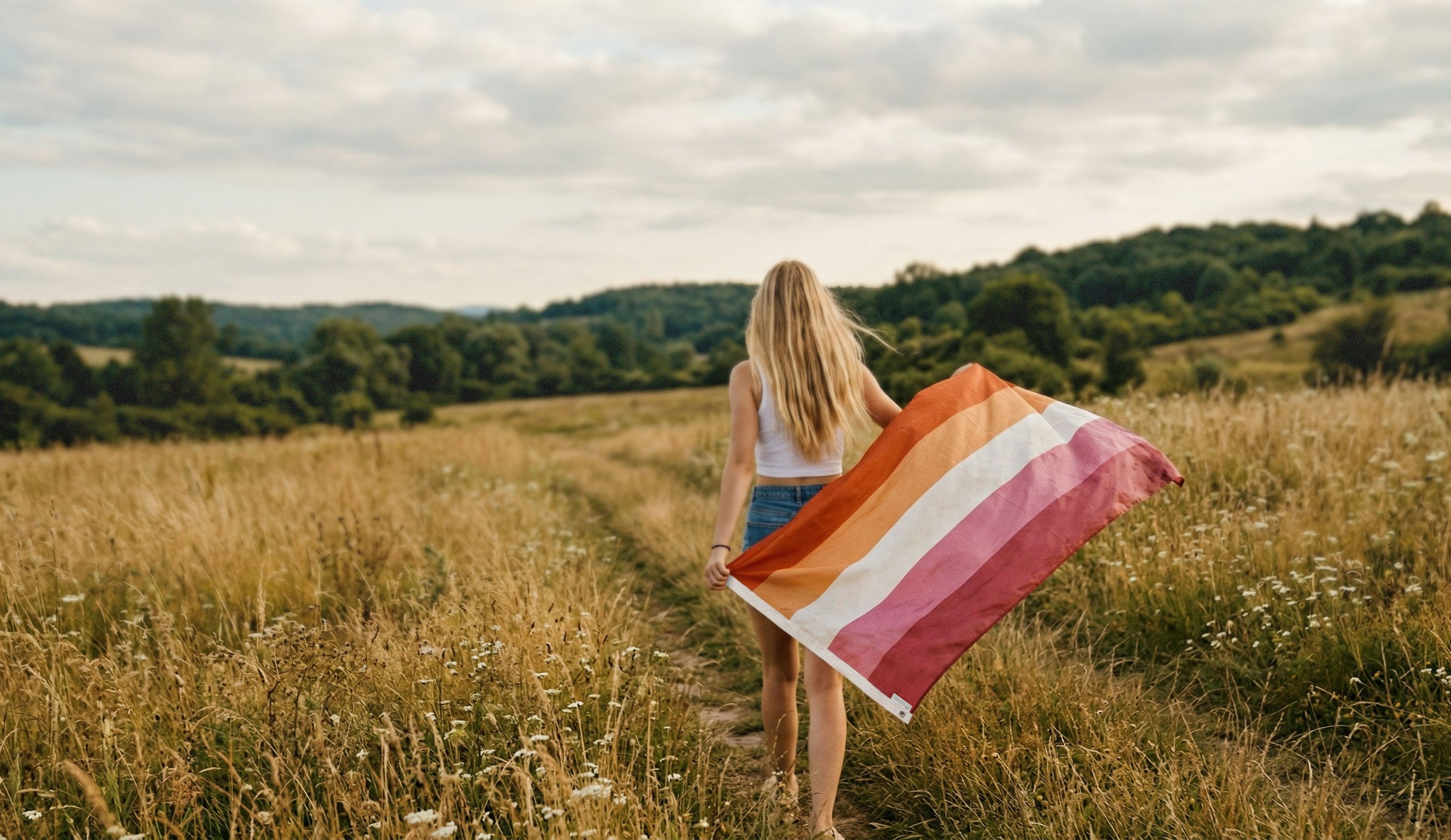 woman holding a Flags For Good lesbian pride  flag in a field walking away from the camera