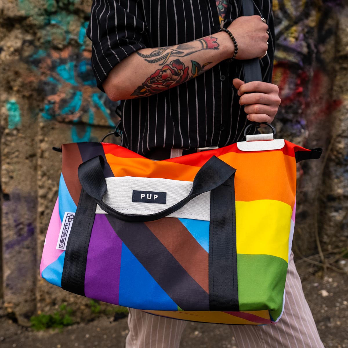 Upcycled Progress Pride Flag Weekender Bag being held by someone in front of graffiti - Flags For Good X PUP