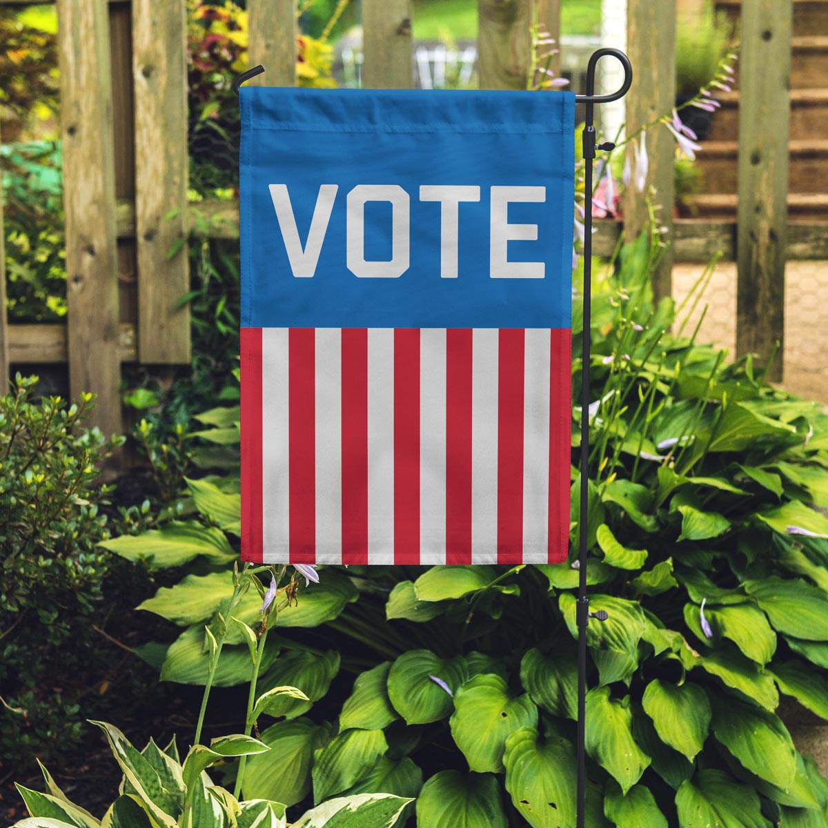 "VOTE" Garden Flag with the word on a blue background and red and white stripes falling vertically down to the bottom in a garden